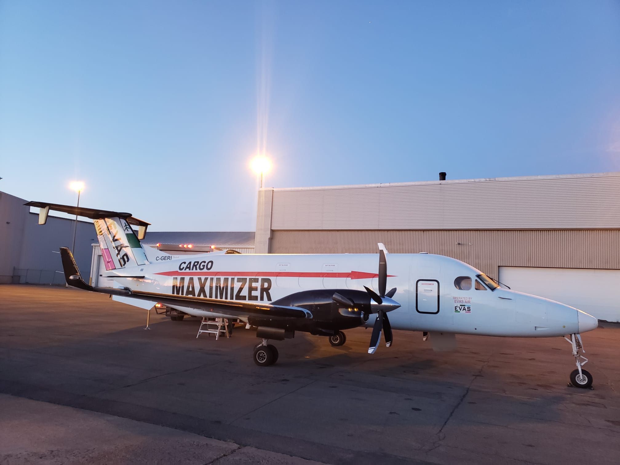 An EVAS Cargo Maximizer Beechcraft 1900D parked on a ramp at dusk beside a hangar.
